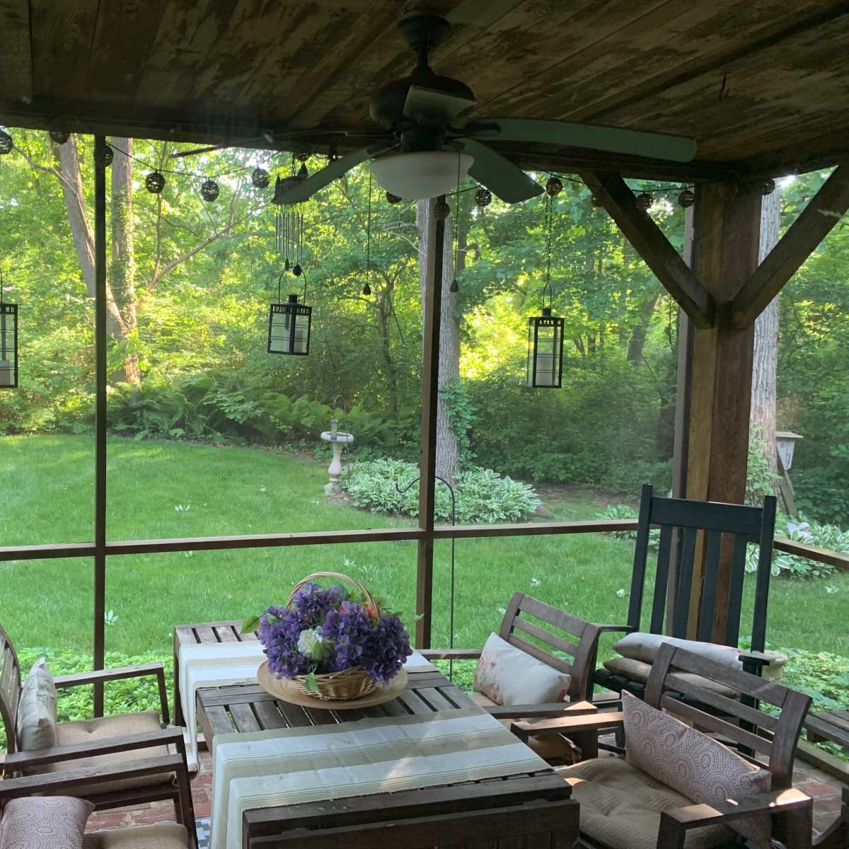A screened porch with wooden furniture, a table adorned with a floral centerpiece, and a view of a lush green garden.