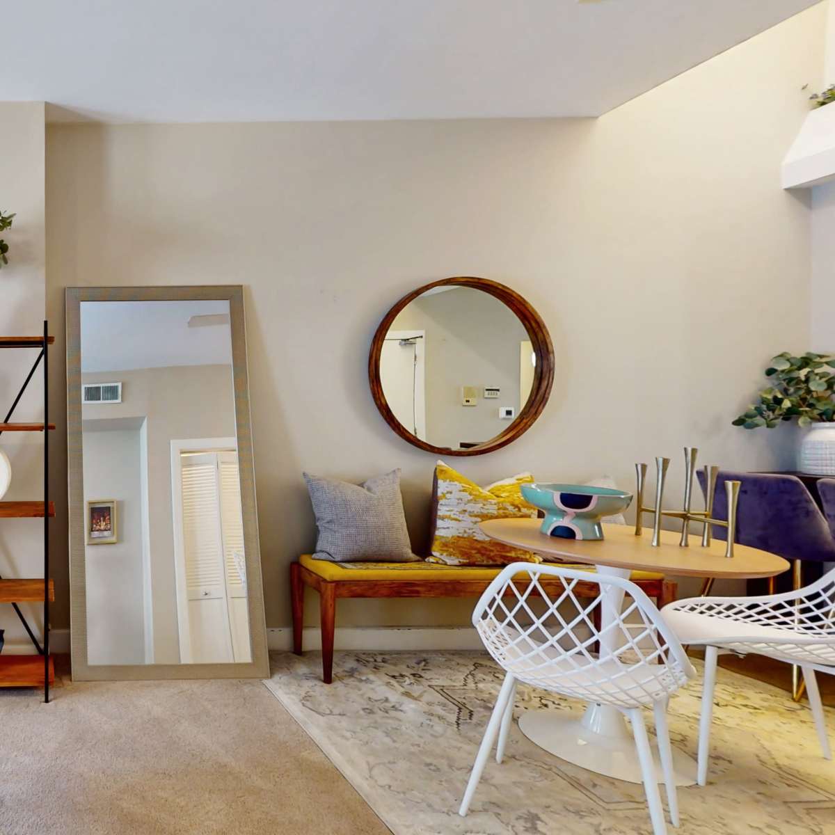 A living room features a cozy seating area with a large mirror, decorative plants, and a round dining table surrounded by chairs.