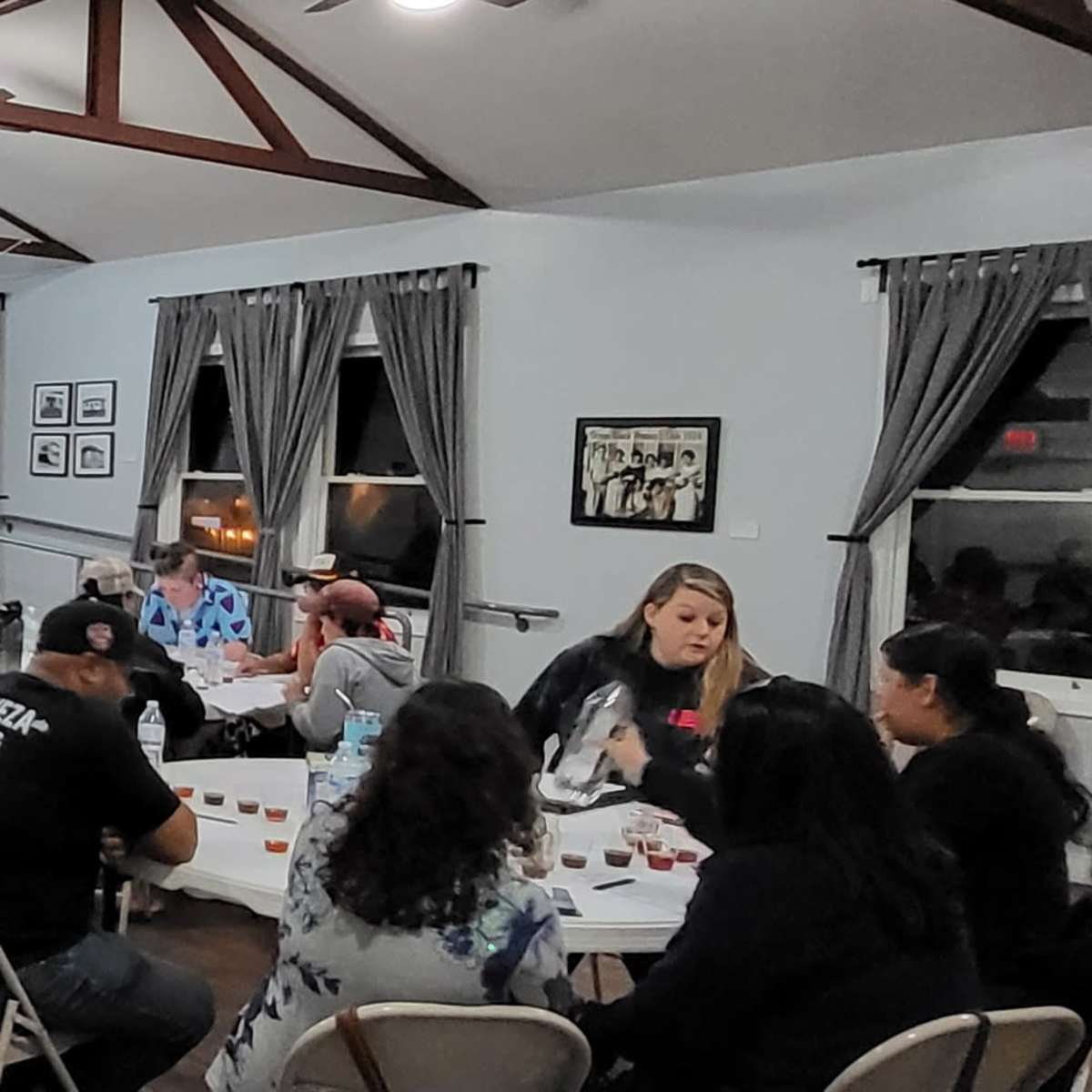 A community gathering takes place in a well-lit hall, with participants seated around several tables, engaging in conversation while a presentation is displayed on a screen in the background.