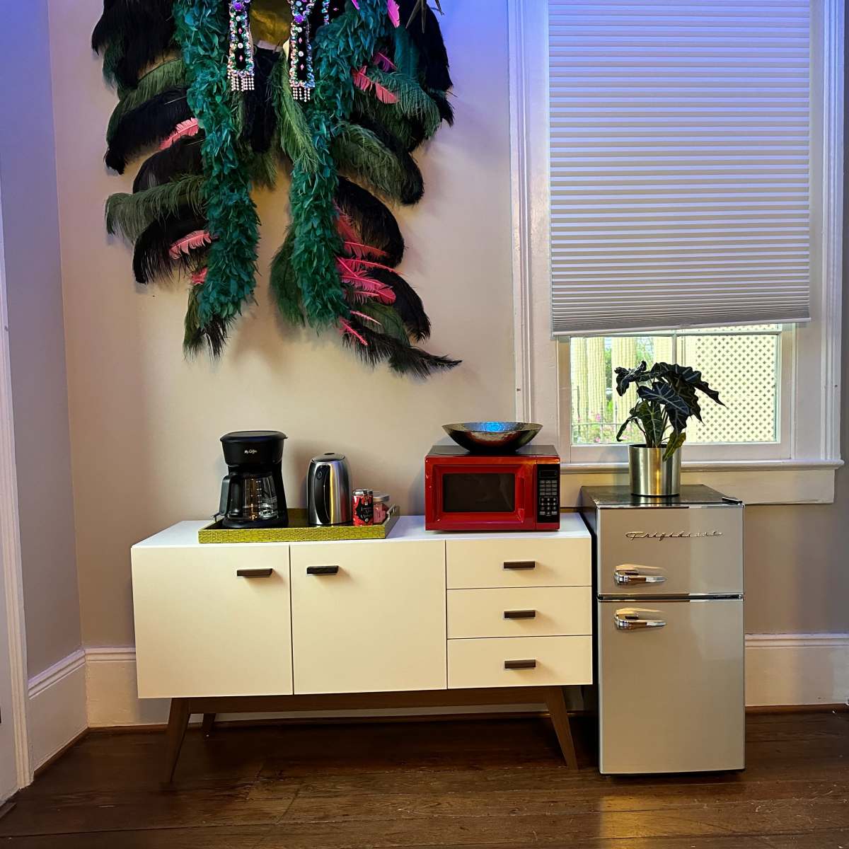 A kitchen corner with a white cabinet, a coffee maker, a red microwave, a decorative bowl, a small silver refrigerator, and a wall adorned with large, colorful feathers.