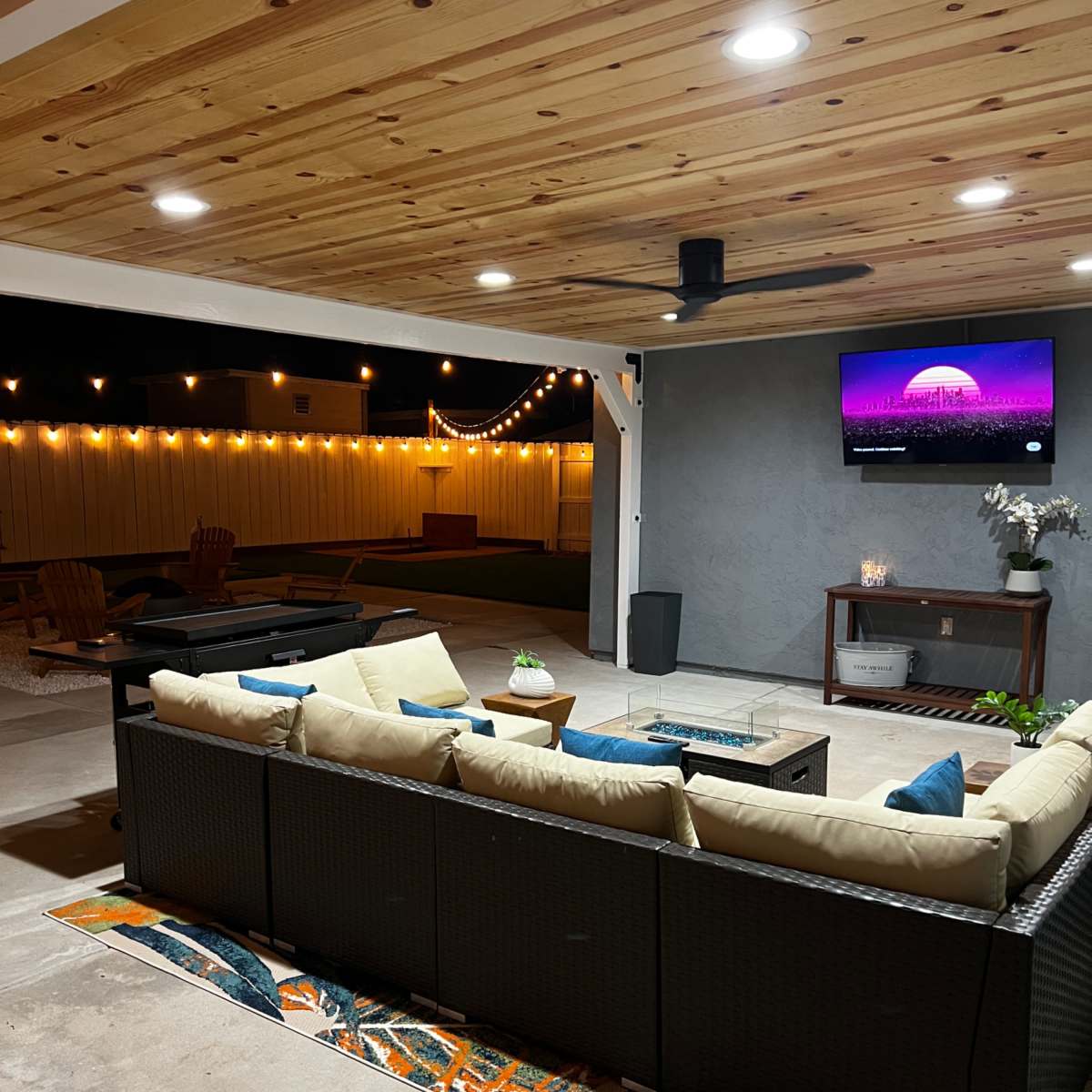 A patio area features a sectional sofa with light-colored cushions, a coffee table, and a wall-mounted TV, illuminated by string lights overhead.
