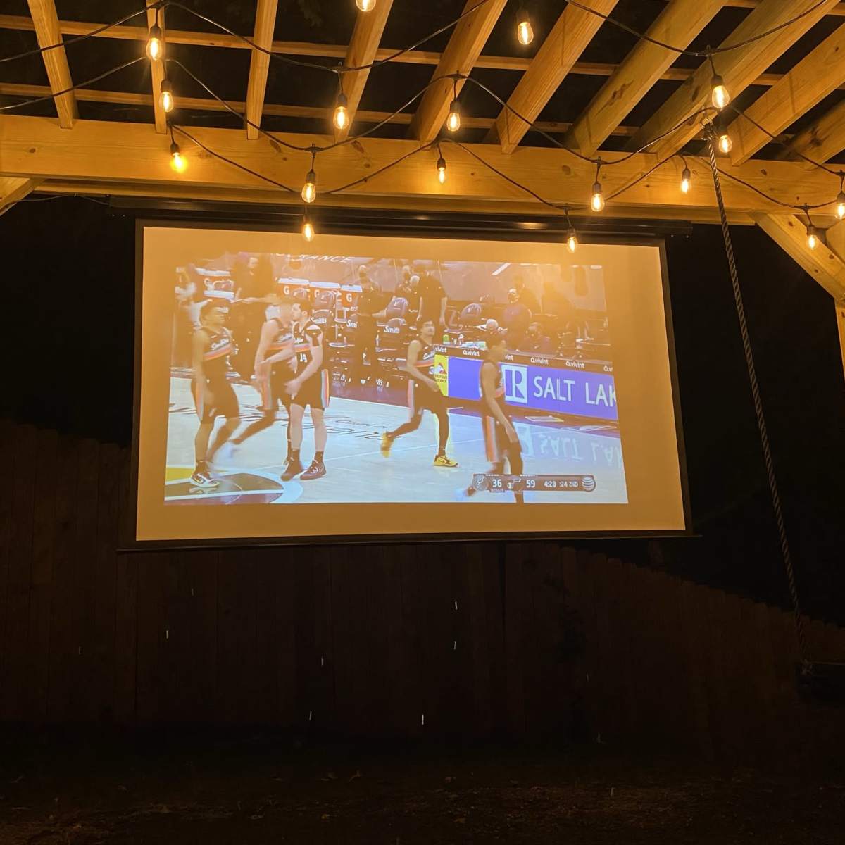 A basketball game is being displayed on a screen in an outdoor pavilion illuminated by string lights.
