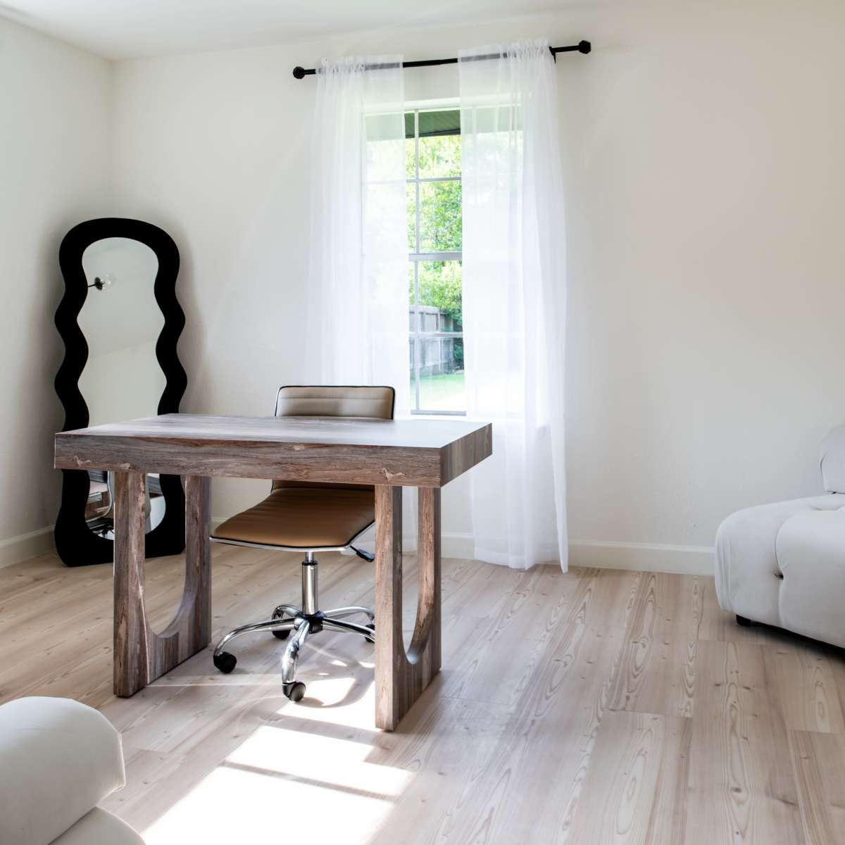 A minimalist office space featuring a wooden desk, an ergonomic chair, and a large mirror beside a window with sheer curtains, all on light-colored wooden flooring.