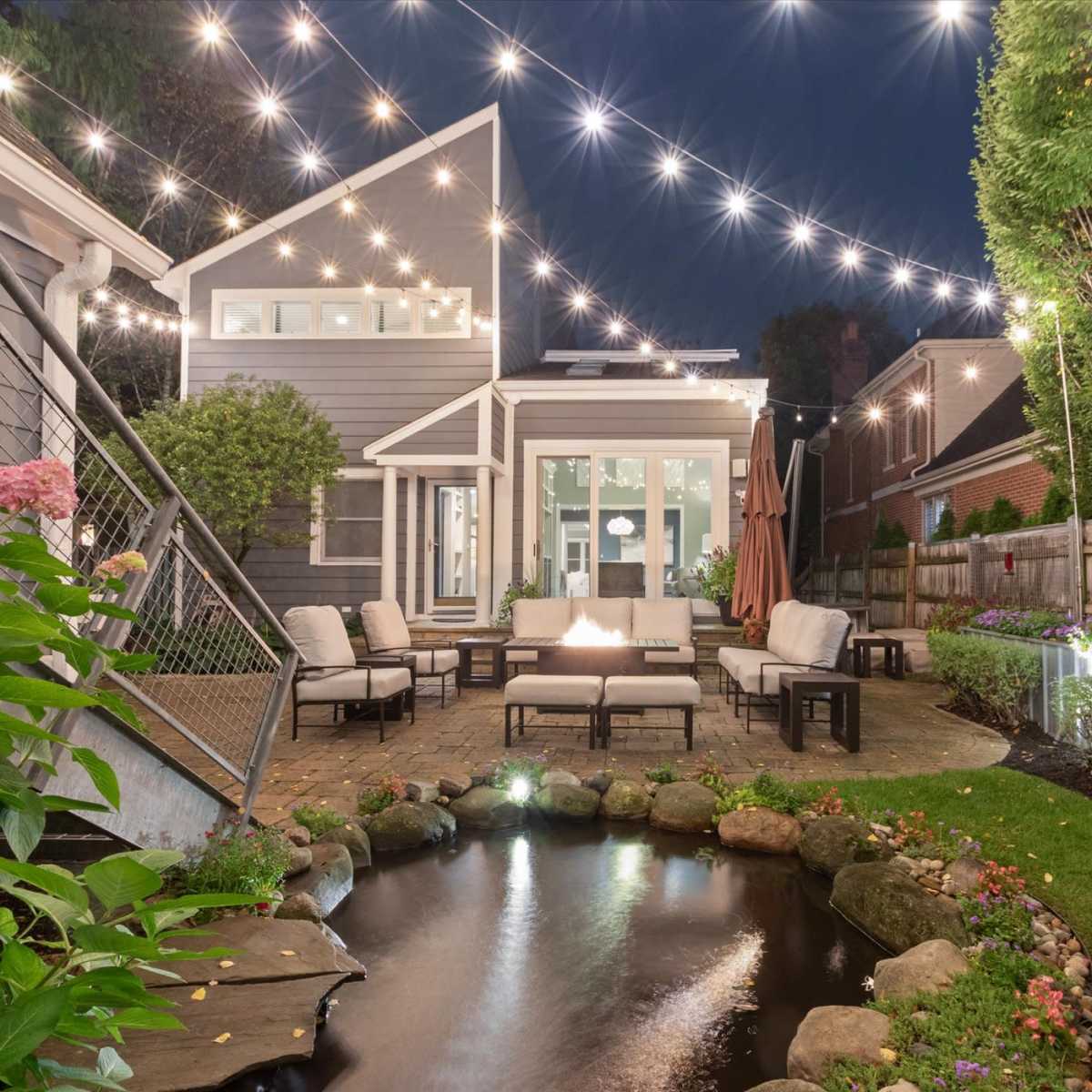 A well-lit backyard features a seating area with a fire pit, surrounded by a pond and neatly arranged landscaping.