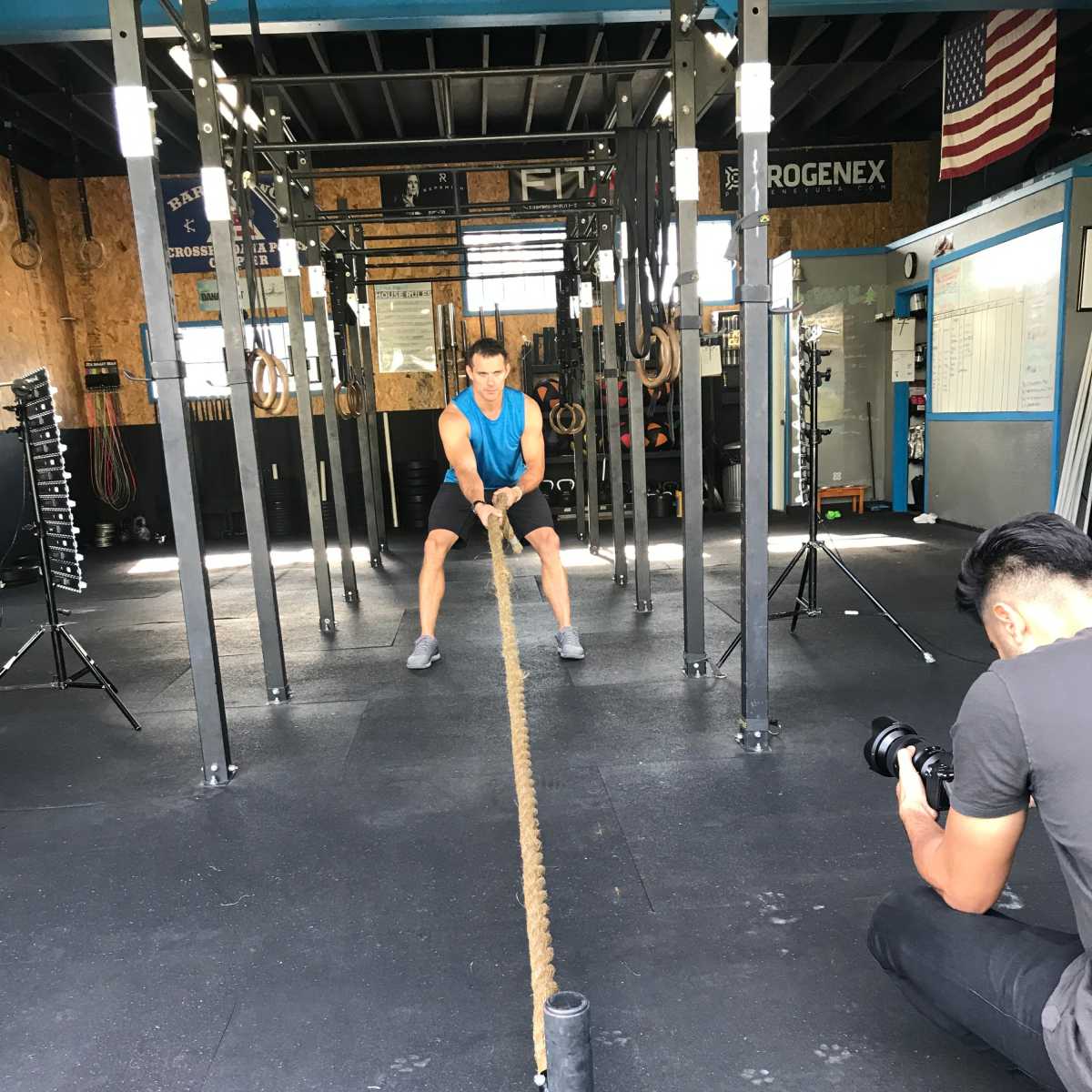 A person is pulling a heavy rope attached to a large weight in a gym while another individual records the activity with a camera.
