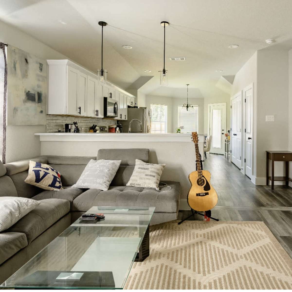 A modern living room with a gray sectional sofa, a glass coffee table, and an acoustic guitar positioned next to it, leading to a hallway and kitchen in the background.