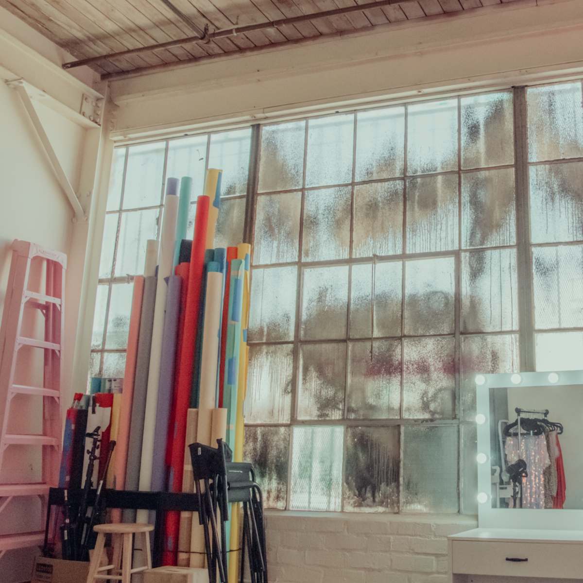 A studio space with large, foggy windows, a mirror with lights, and colorful rolls of material stacked against the wall.