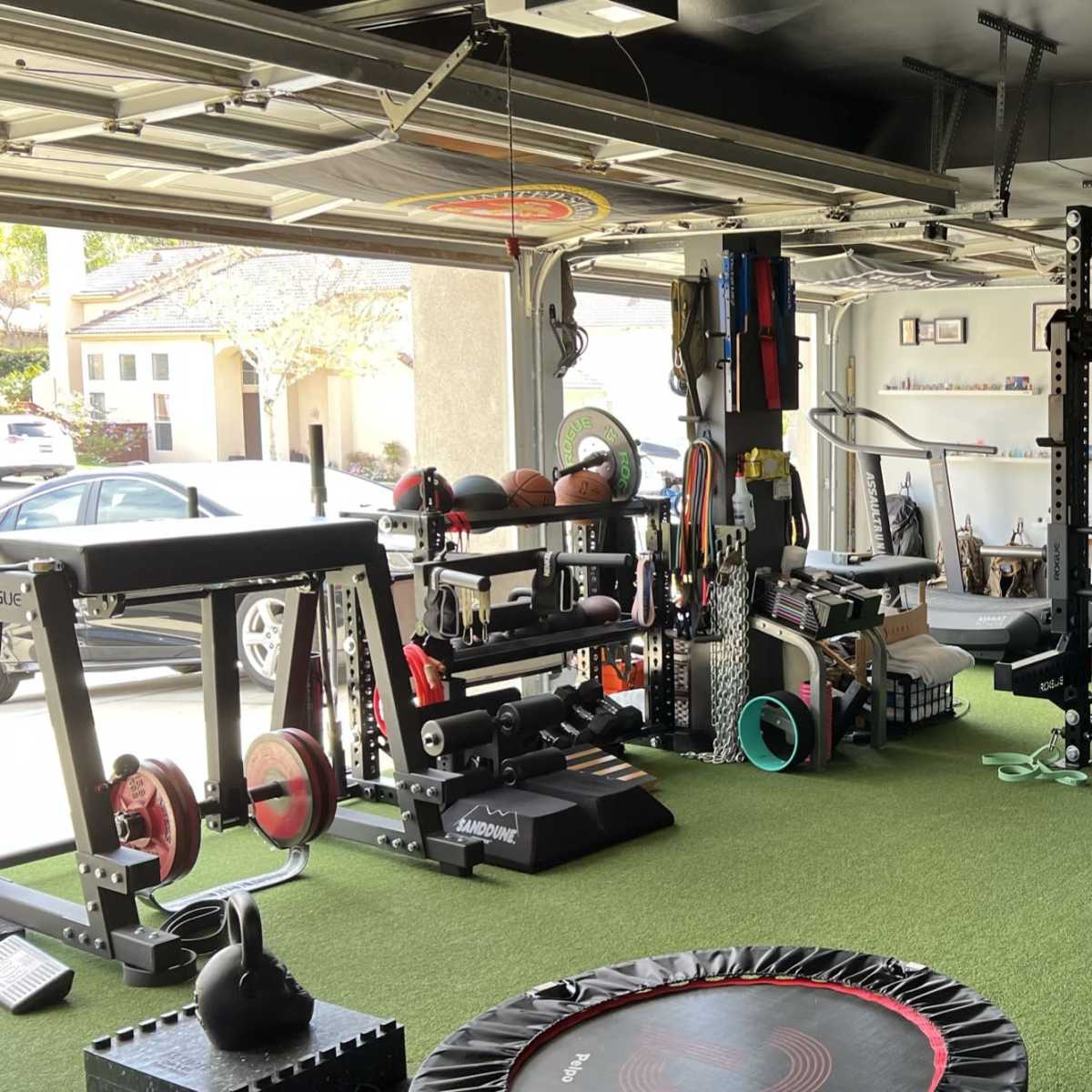 A home garage gym equipped with various fitness machines, weights, and exercise accessories on a green turf floor.