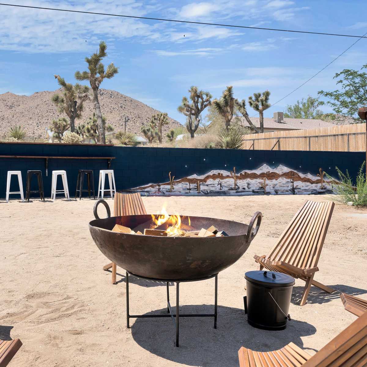 A circular fire pit surrounded by wooden chairs sits in a sandy area with a mural and desert landscape in the background.