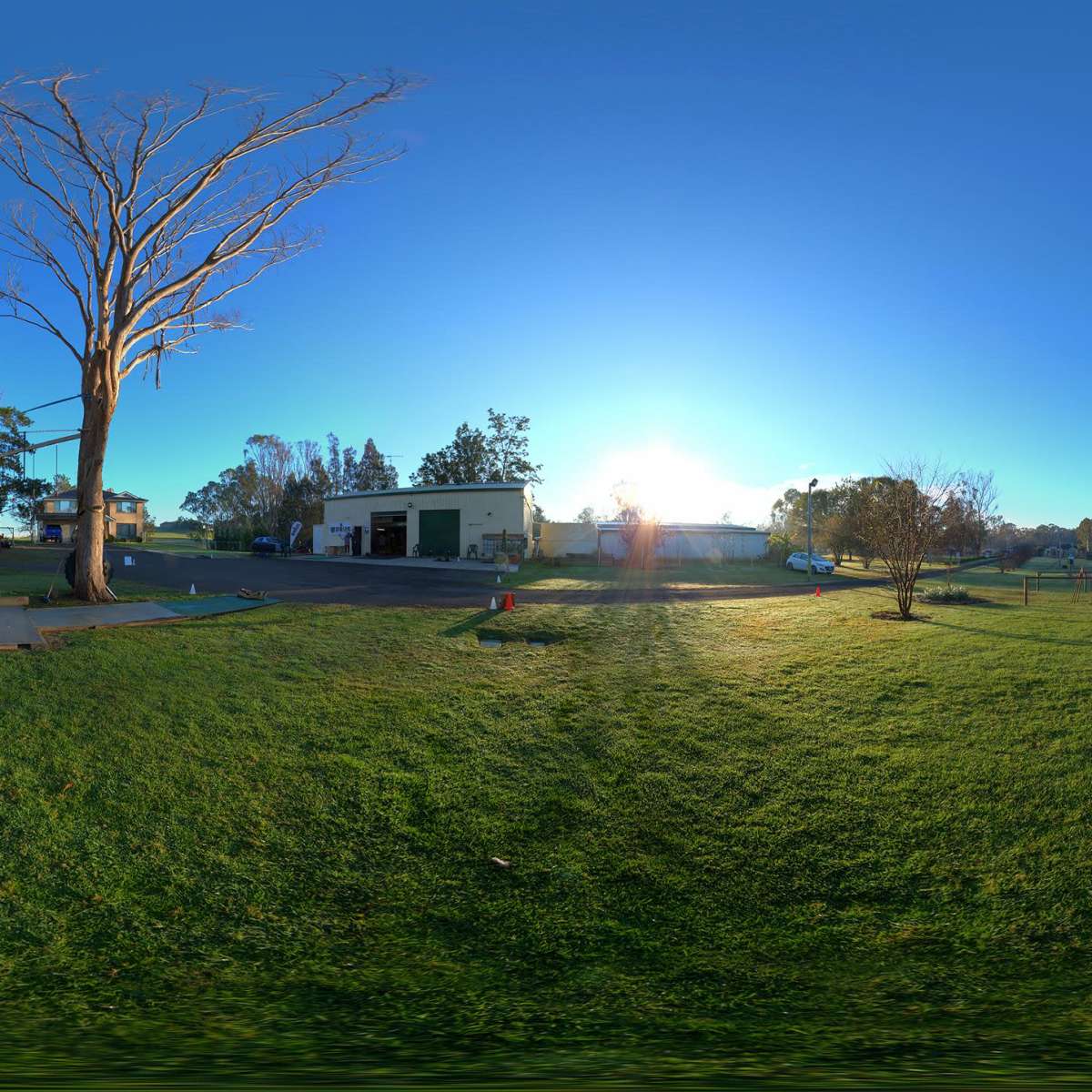 A panoramic view of a park-like area with a grass field, a half-pipe structure, and surrounded by trees and buildings under a clear blue sky.