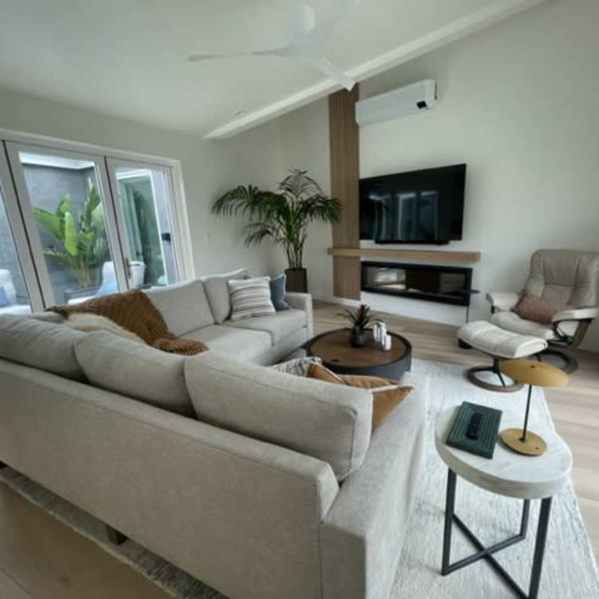 A modern living room features a light-colored sectional sofa, a round coffee table, a wall-mounted television, and a cozy armchair, with large windows allowing natural light to fill the space.