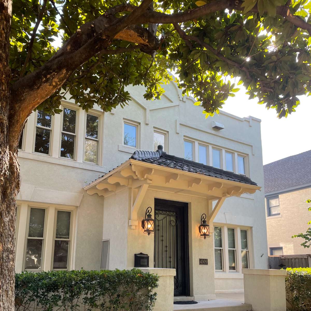 A two-story, light-colored house with decorative facade details, flanked by a garden and large tree.