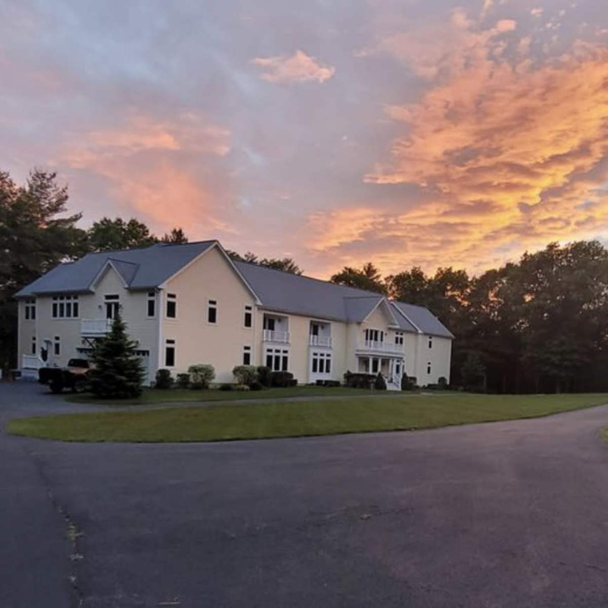 A large, two-story house with a manicured lawn is situated beside a winding driveway under a colorful sunset sky.