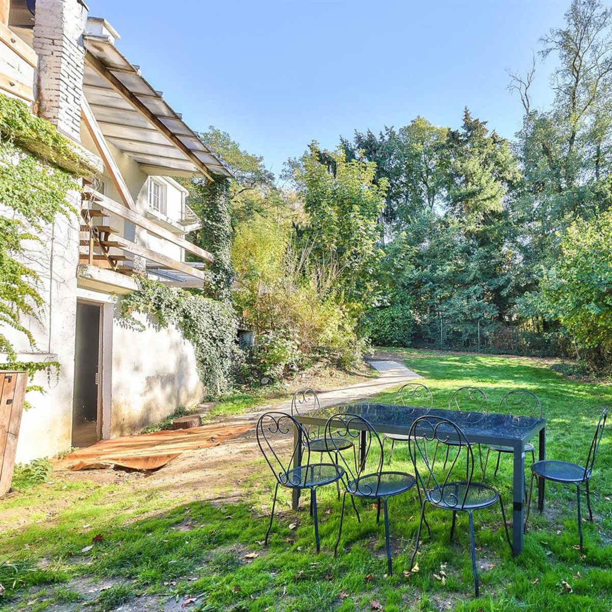 A small black table with six chairs sits on a grassy area beside a house, surrounded by trees and vegetation.
