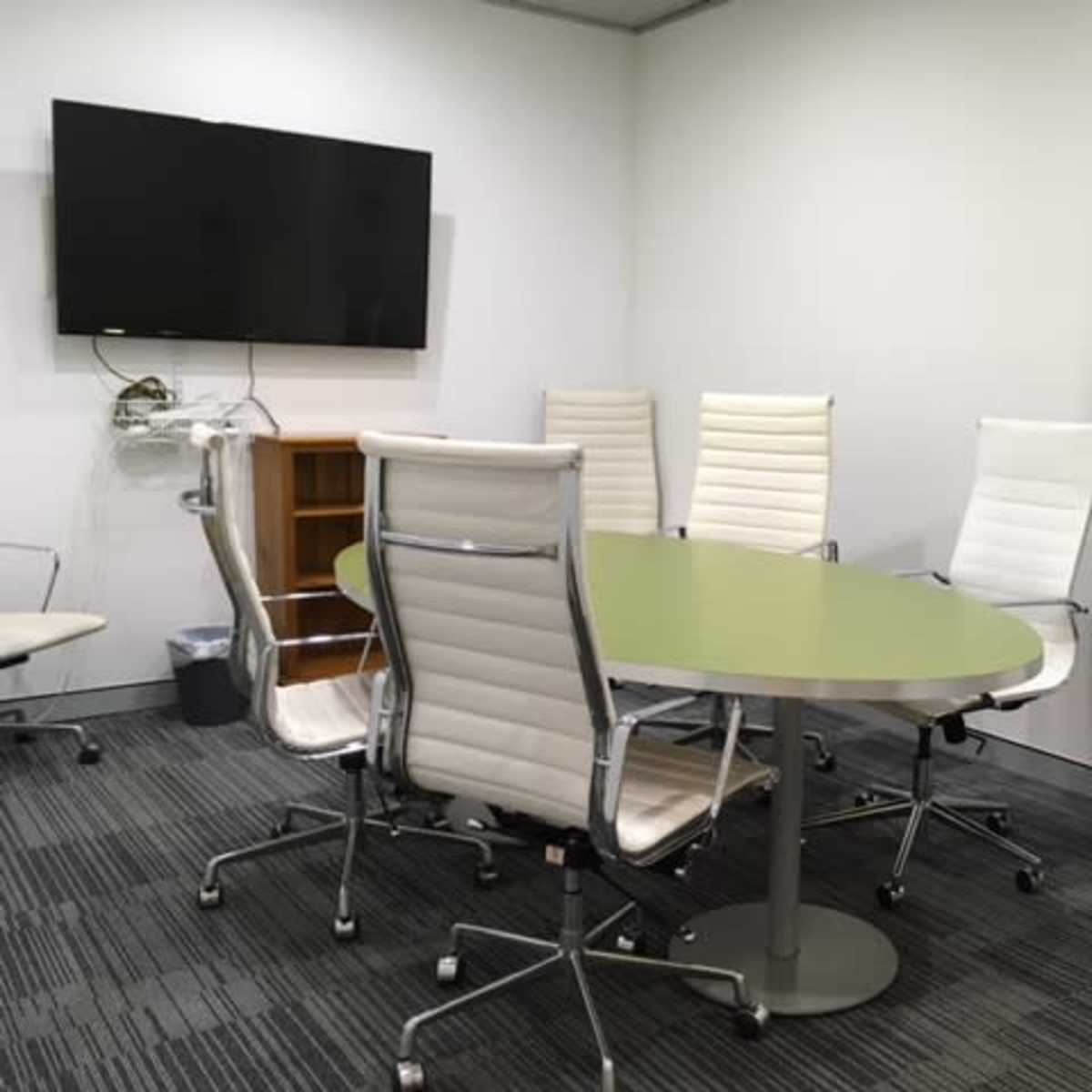 A conference room features a green oval table surrounded by six white ergonomic chairs and a wall-mounted television.