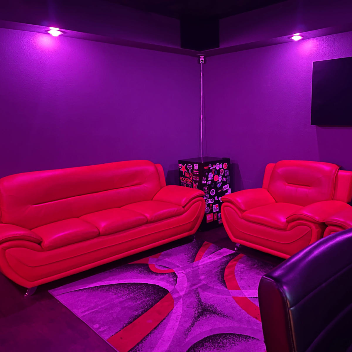 A room with two red sofas, a dark purple wall, and a television mounted on the wall, illuminated by purple lighting.