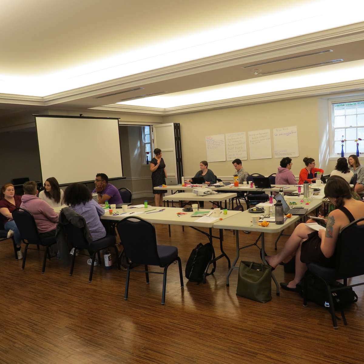 A group of people is seated at tables in a classroom setting, actively engaged in a workshop with a presentation screen and notes displayed on the walls.