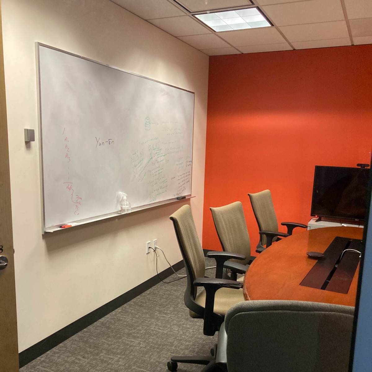 A small conference room with a round table, five chairs, a whiteboard on one wall, and a television screen.