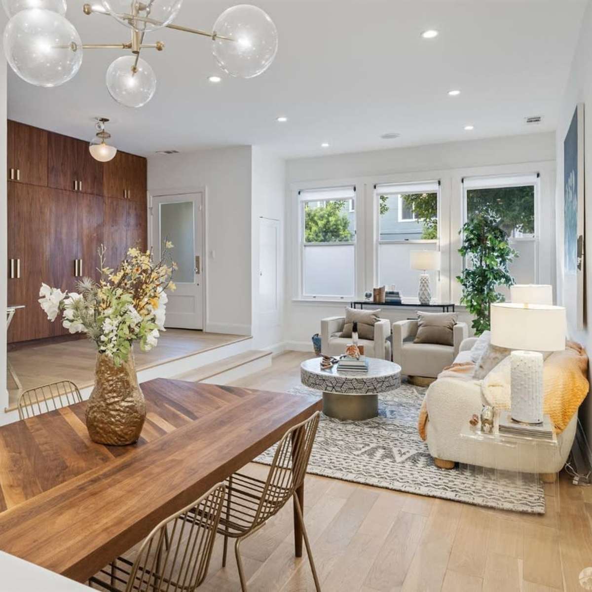 A modern living space featuring a dining area with a wood table, a bright sitting area with a couch and coffee table, and a kitchen with wooden cabinetry.