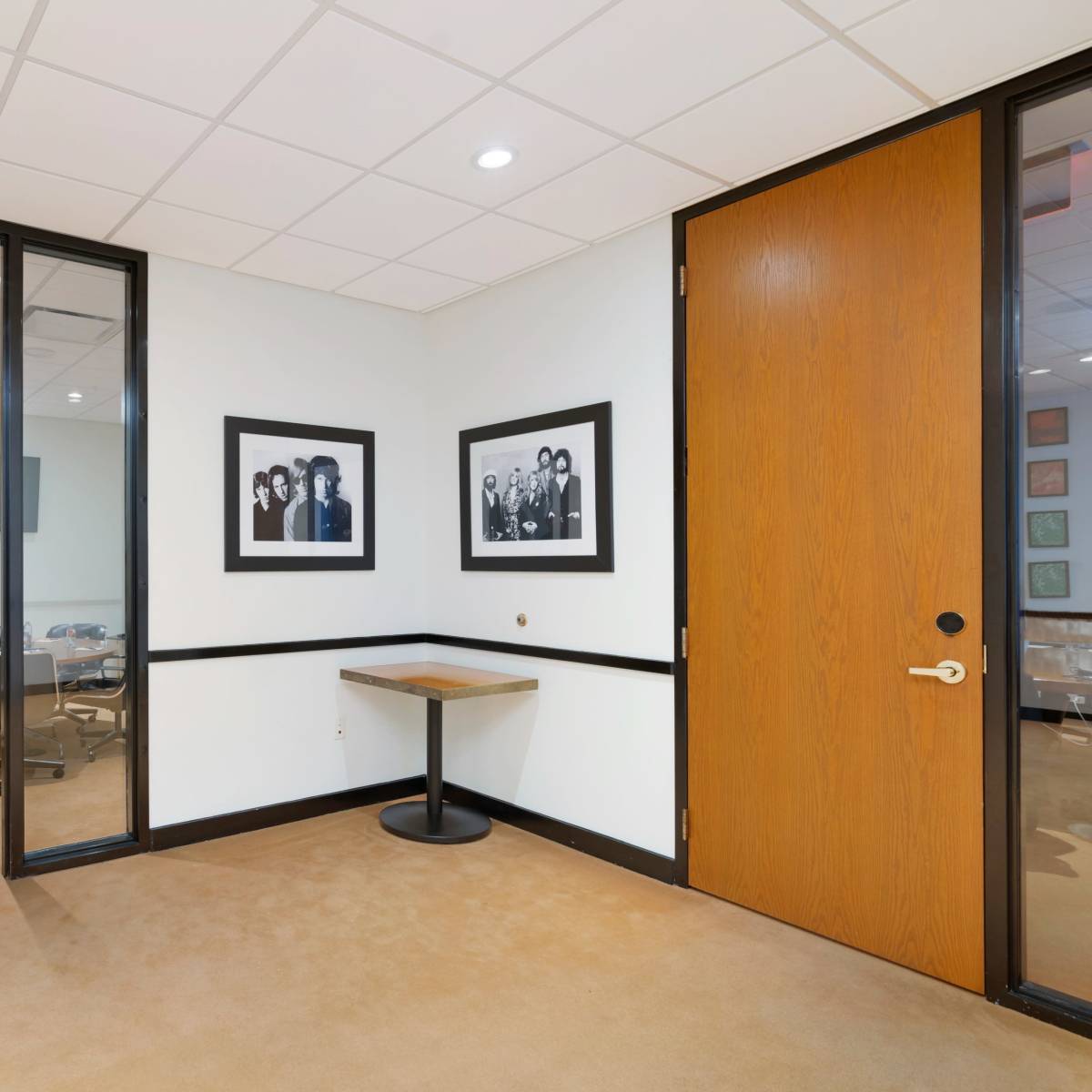 An office hallway with a glass-enclosed meeting room, a wooden door, and two framed black-and-white photographs on the wall.