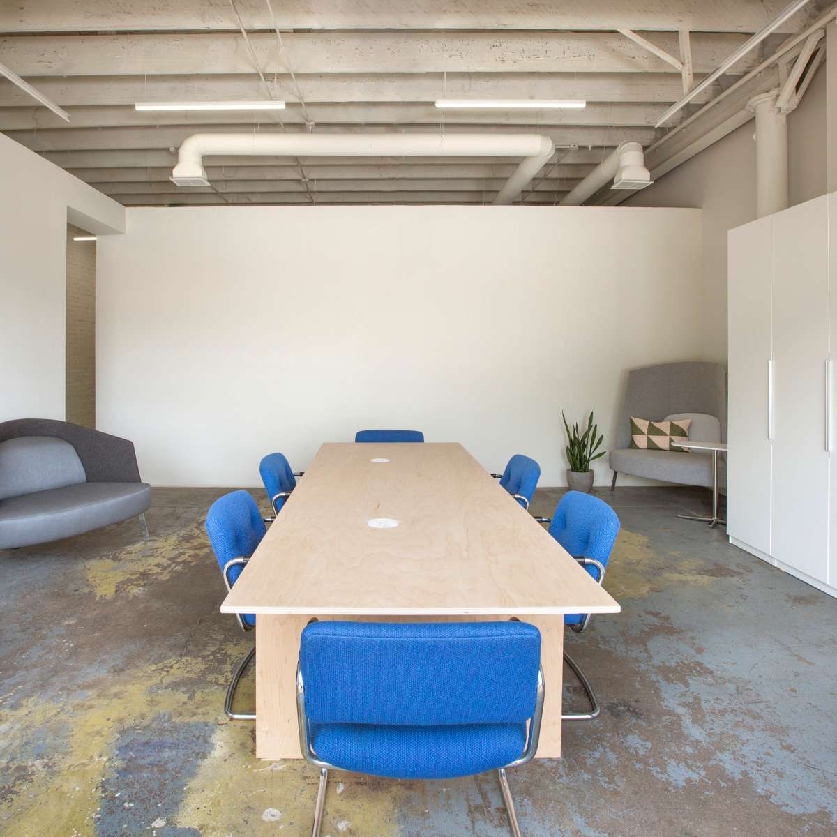 A large wooden table is surrounded by eight blue chairs in a minimalistic meeting room with gray furnishings and a potted plant.