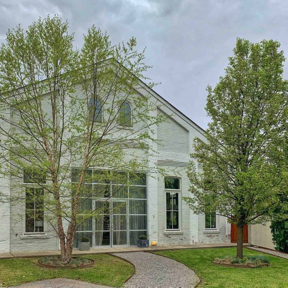 A white brick house with large windows and two trees flanking a stone pathway leads to the entrance.