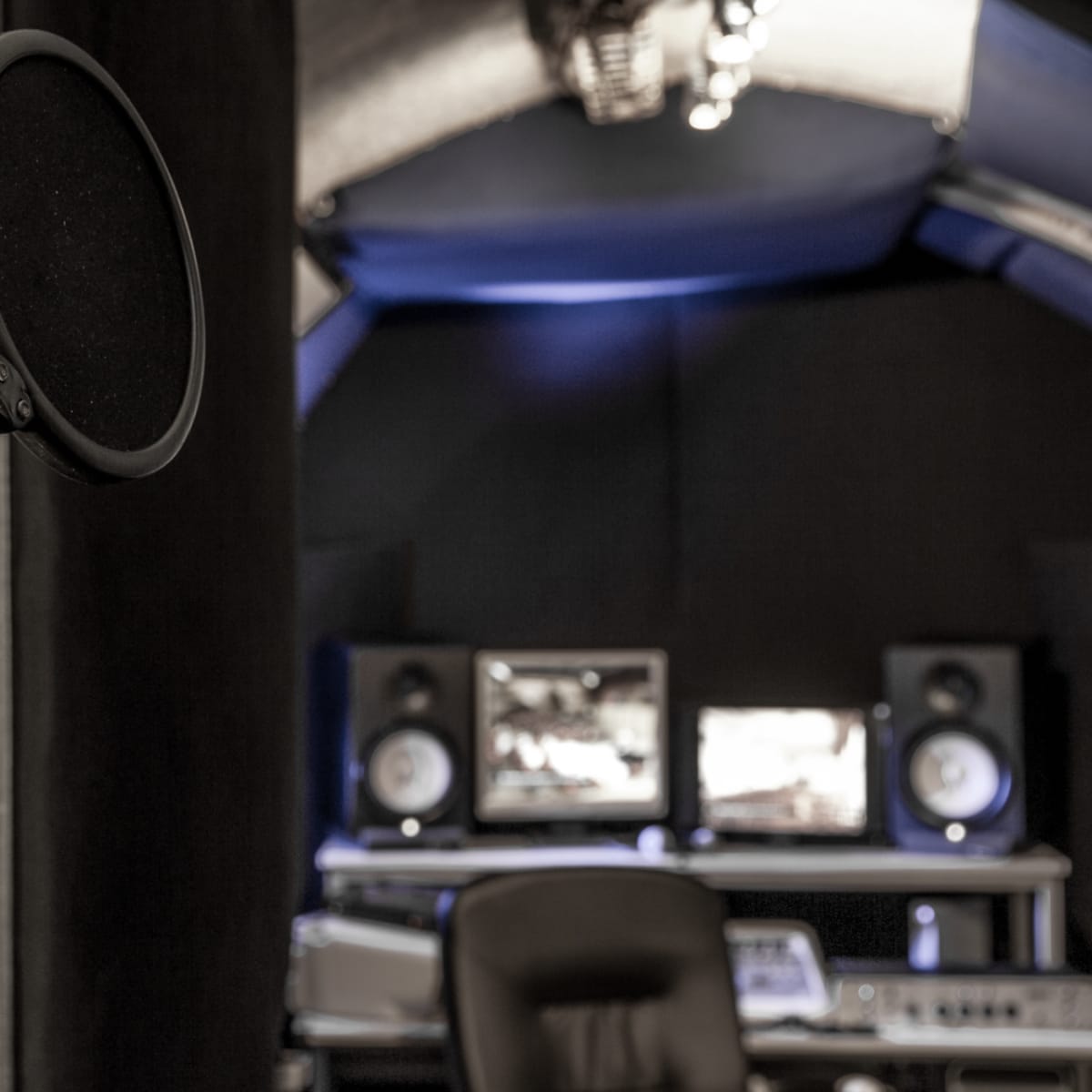 A microphone with a pop filter stands in the foreground of a sound recording studio, featuring computer monitors and audio equipment in the background.