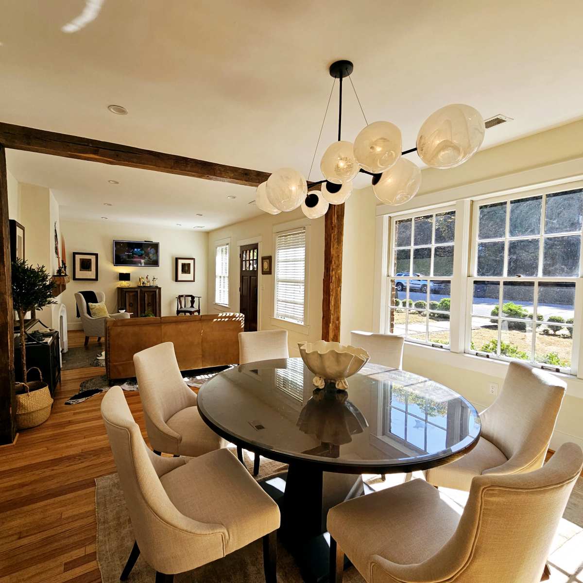A round dining table with eight chairs is positioned in a well-lit room featuring large windows and a chandelier.