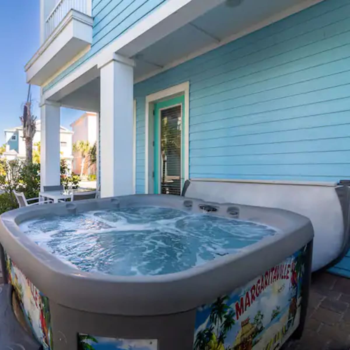 A hot tub filled with water sits beside a patio area with lounge chairs and palm trees in a residential setting.