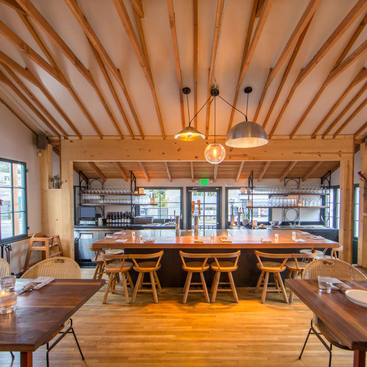 A modern restaurant interior with wooden beams, a central bar area, and tables set for dining.