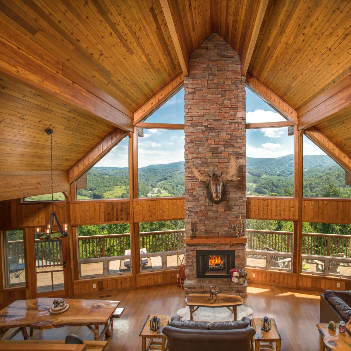 A spacious living room with a high wooden ceiling, featuring a central stone fireplace and large windows that offer a view of the surrounding hills.