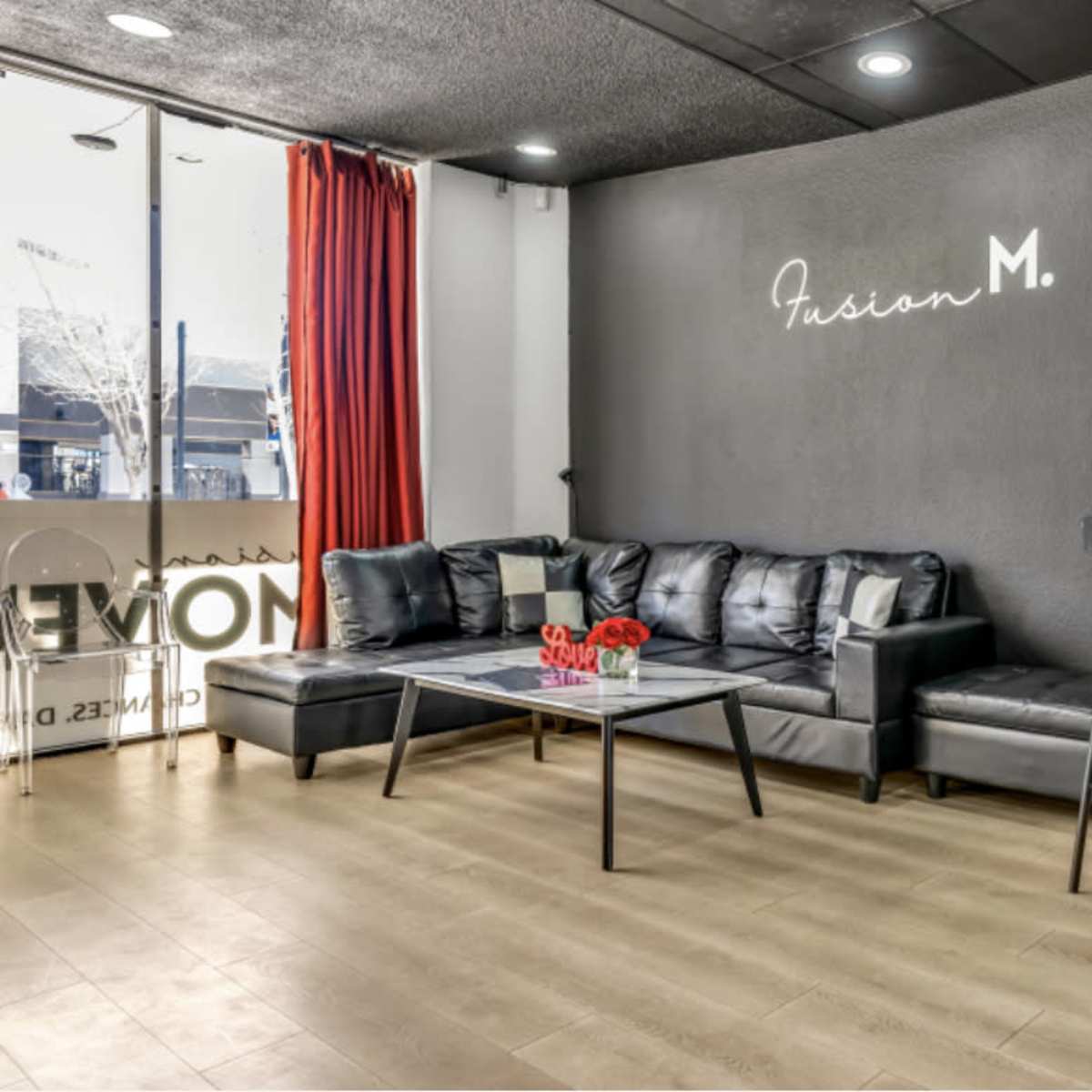 A modern lounge area with a black leather couch, a glass coffee table, and bar stools against a dark wall featuring the name "Fusion M."