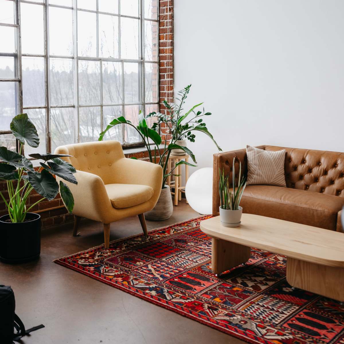 A cozy living room features a tan leather sofa, a yellow armchair, a wooden coffee table, a colorful rug, and large windows surrounded by plants.