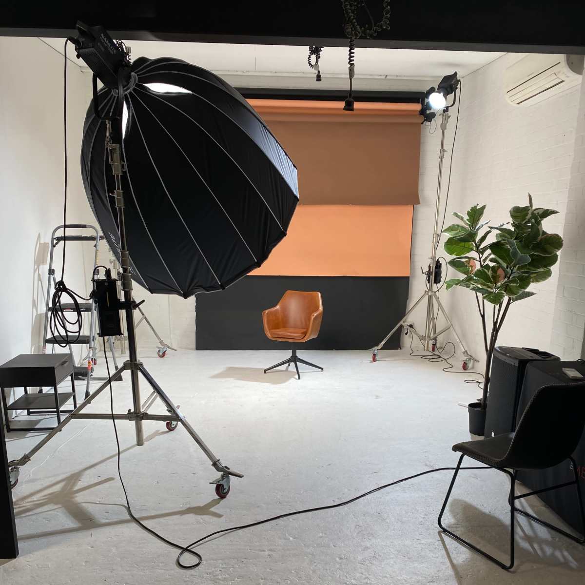 A photography studio features a large umbrella light, an empty orange-backed set, a brown chair, and various equipment.
