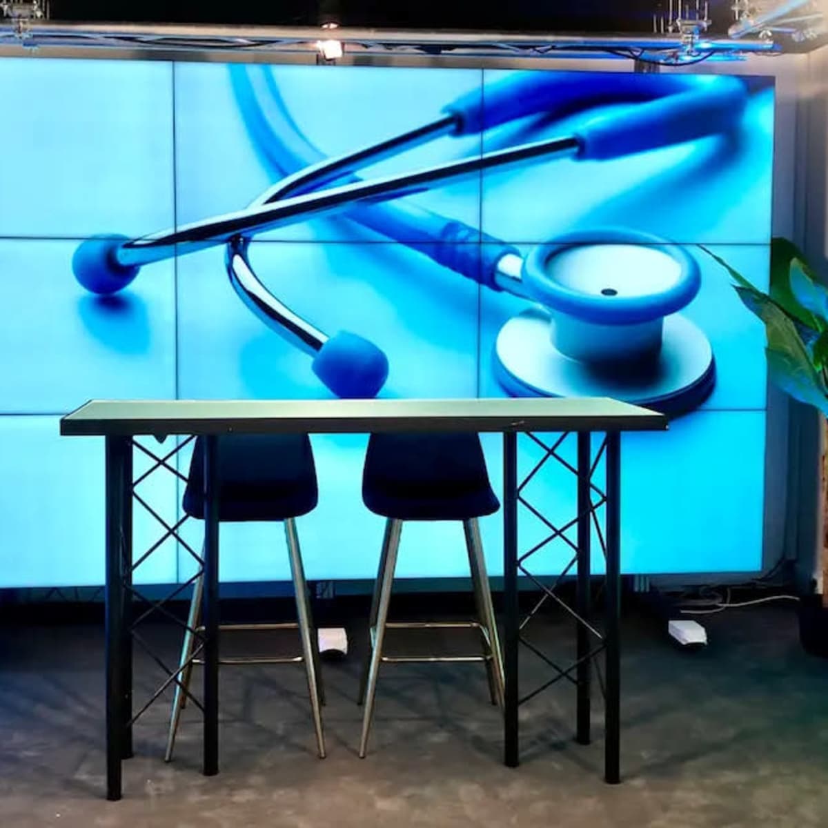 A stage setting with a large screen displaying a stethoscope, two high stools, and green plants in black shelving units.
