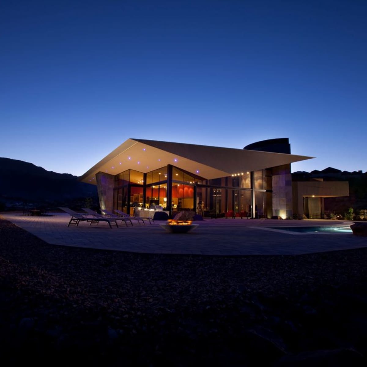 A modern villa with large glass windows and a distinctive roof design is illuminated at dusk, surrounded by landscaping and a swimming pool.