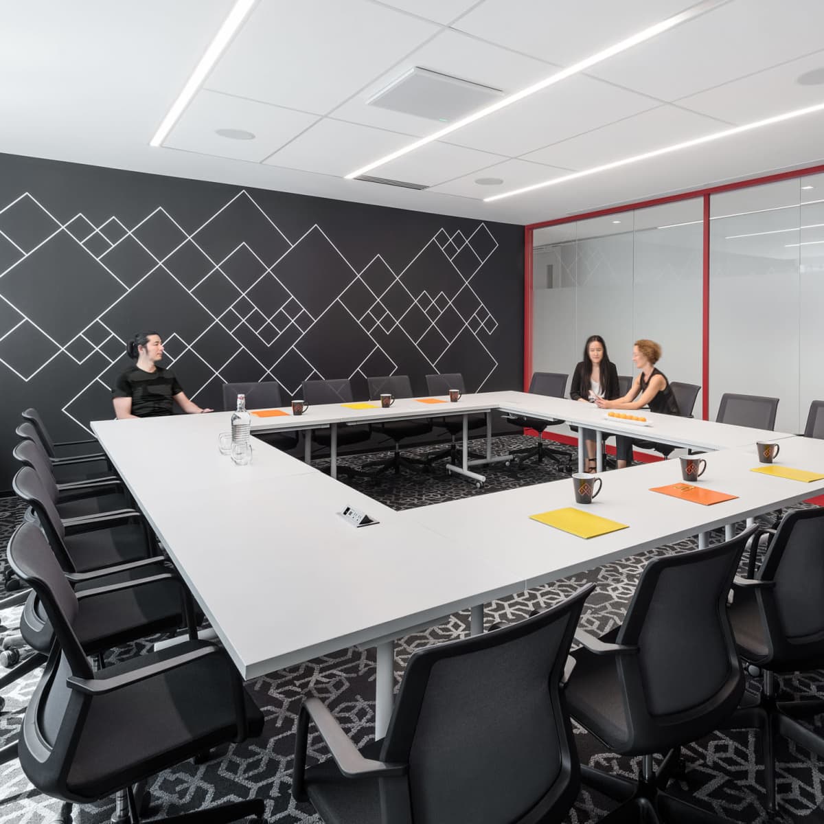A modern conference room features a large rectangular table surrounded by black chairs, with two women seated at one end and geometric patterns on the walls.