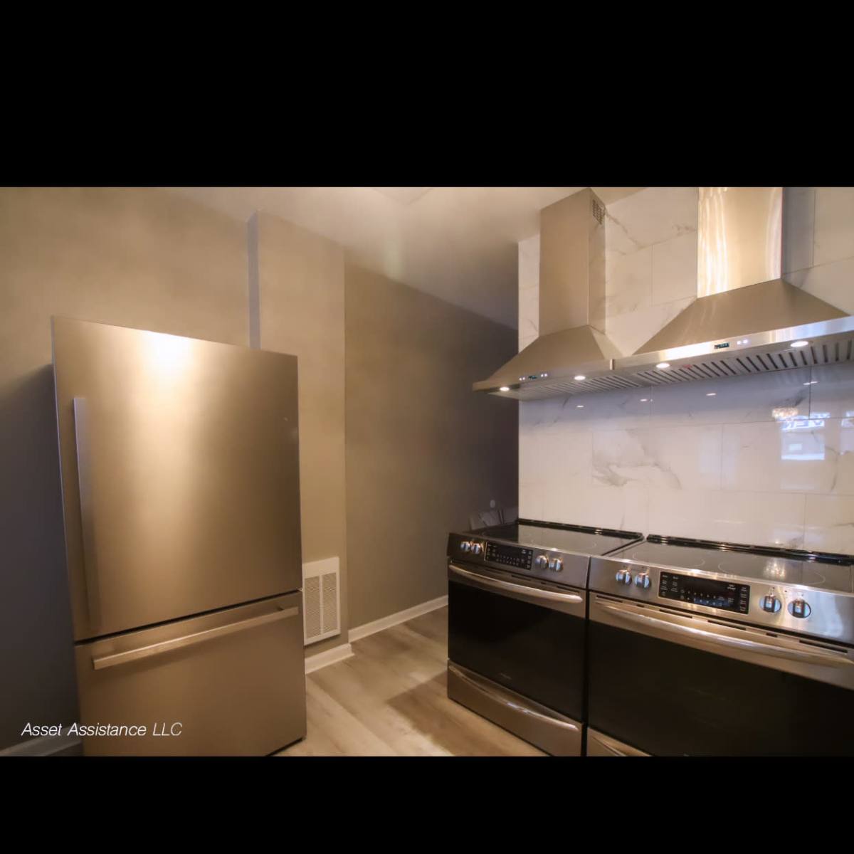 A modern kitchen with a stainless steel refrigerator and a black oven range installed next to a sleek exhaust hood and a neutral gray wall.