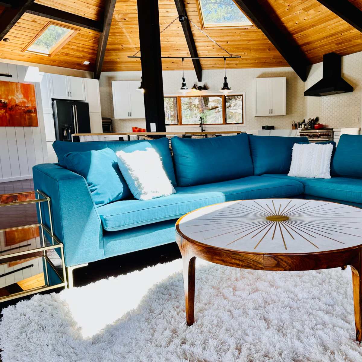 A teal sectional sofa with a white throw pillow is positioned next to a round coffee table on a plush rug in a bright, airy living space with wooden beams and large windows.