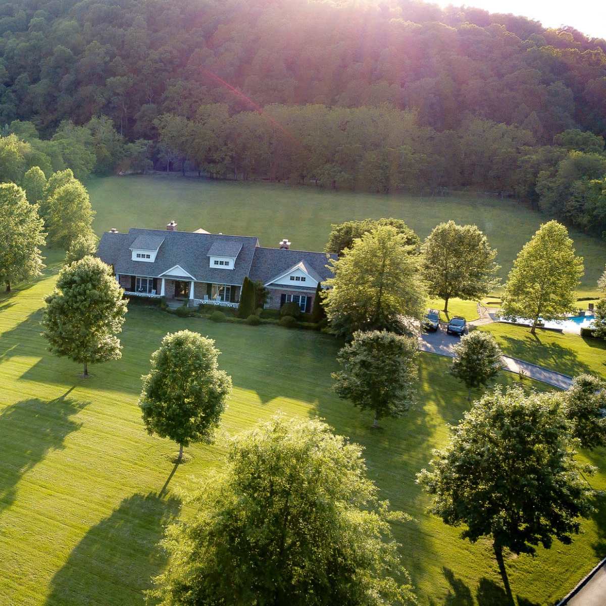 A large house is surrounded by a lush lawn and trees, set against a backdrop of rolling hills.