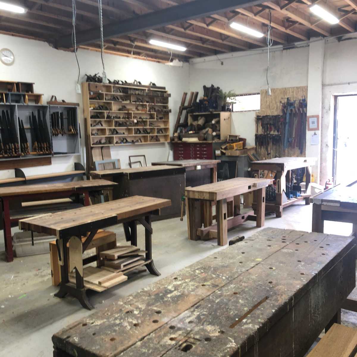 A woodworking shop with multiple workbenches, tools neatly arranged on shelves, and various woodworking equipment visible throughout the space.