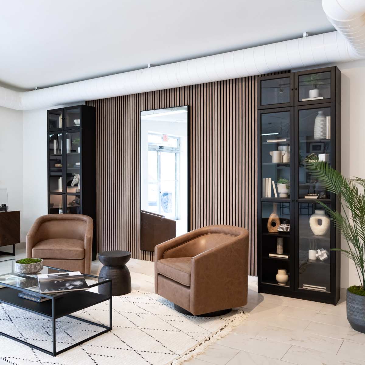 A modern living room set with two brown leather chairs, a glass coffee table, a patterned rug, and shelves displaying decorative items against a wall with vertical wood paneling.