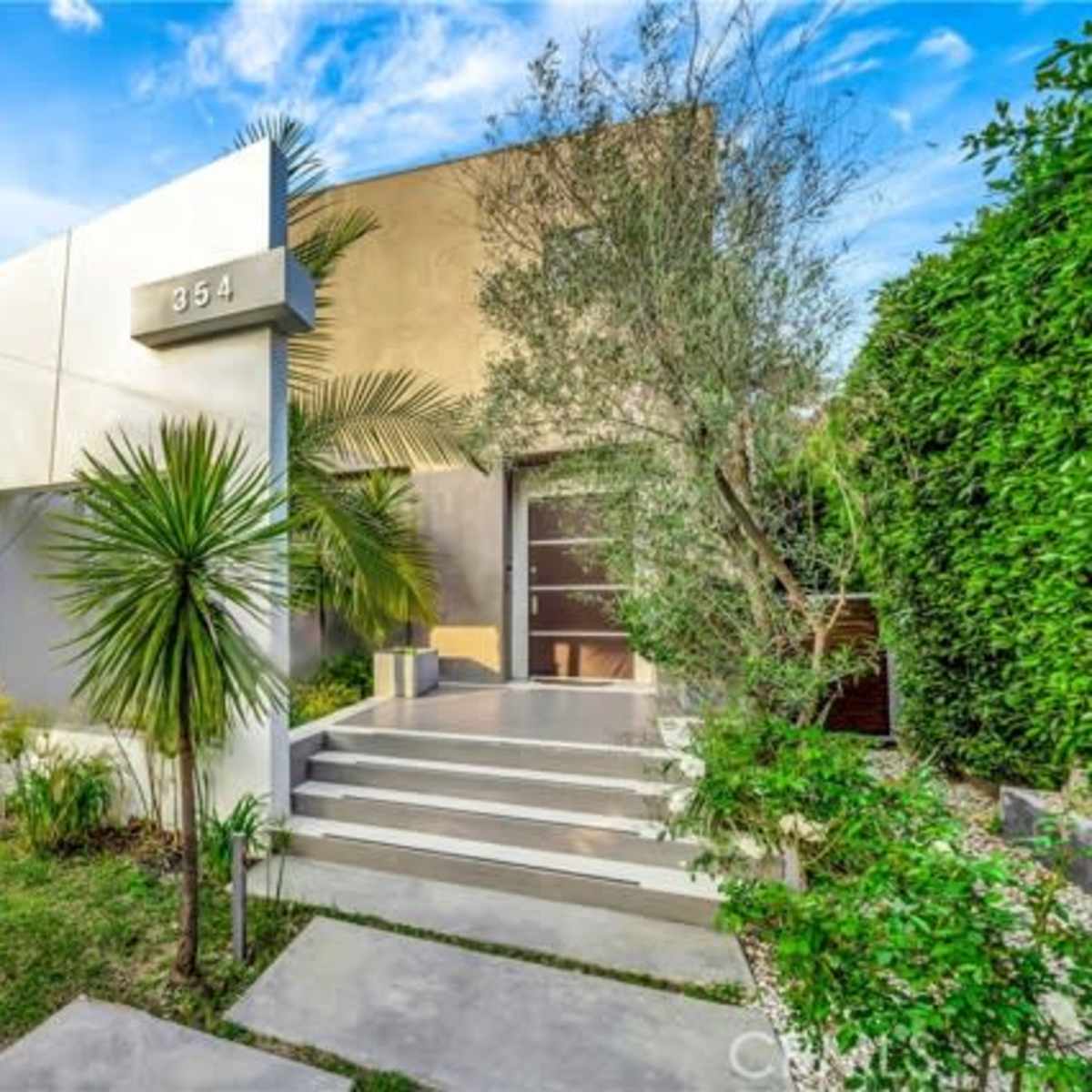 A modern house with a geometric design, featuring a concrete facade, palm trees, and a pathway leading to the entrance.