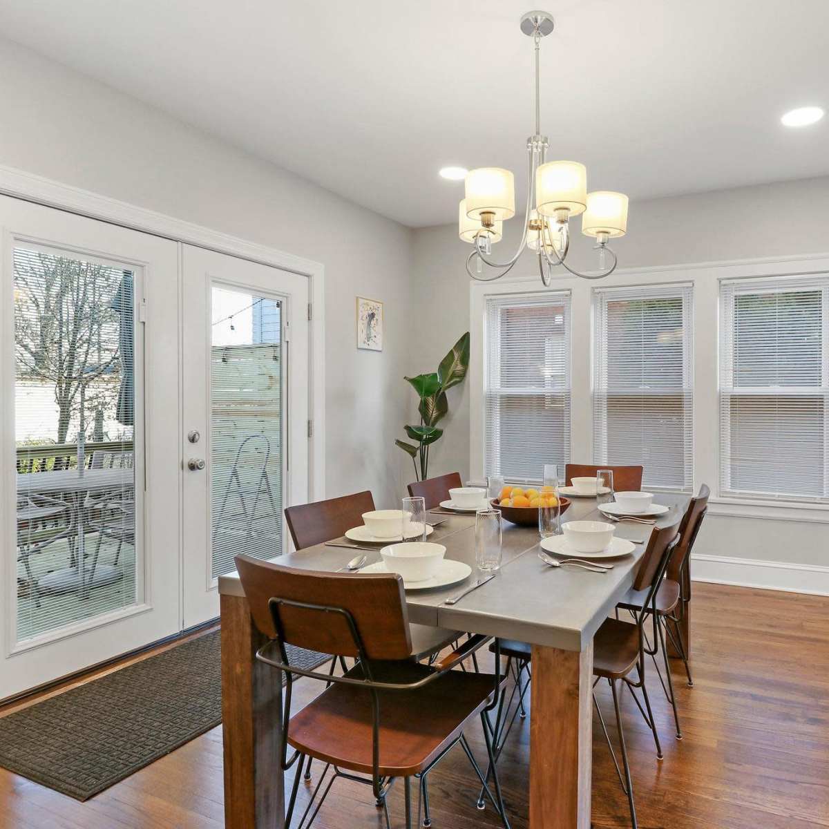 A dining area features a table set for dinner with wooden chairs, a chandelier overhead, and large doors leading to a patio.