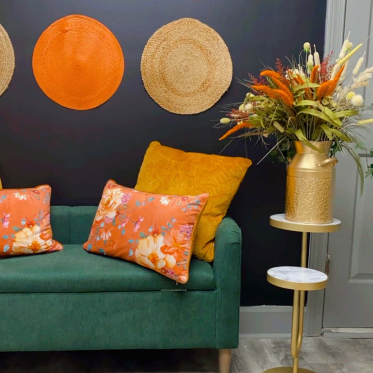 A green sofa with patterned orange and yellow cushions is positioned against a dark wall adorned with woven circular wall art, next to a gold side table topped with a floral arrangement.