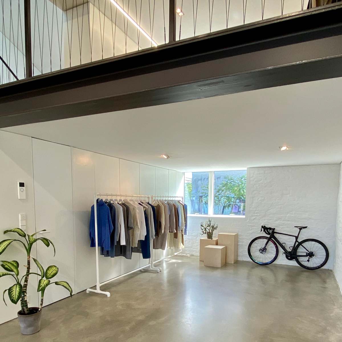 A minimalistic interior space featuring a clothing rack with various garments, a potted plant, a bicycle, and a window allowing natural light.