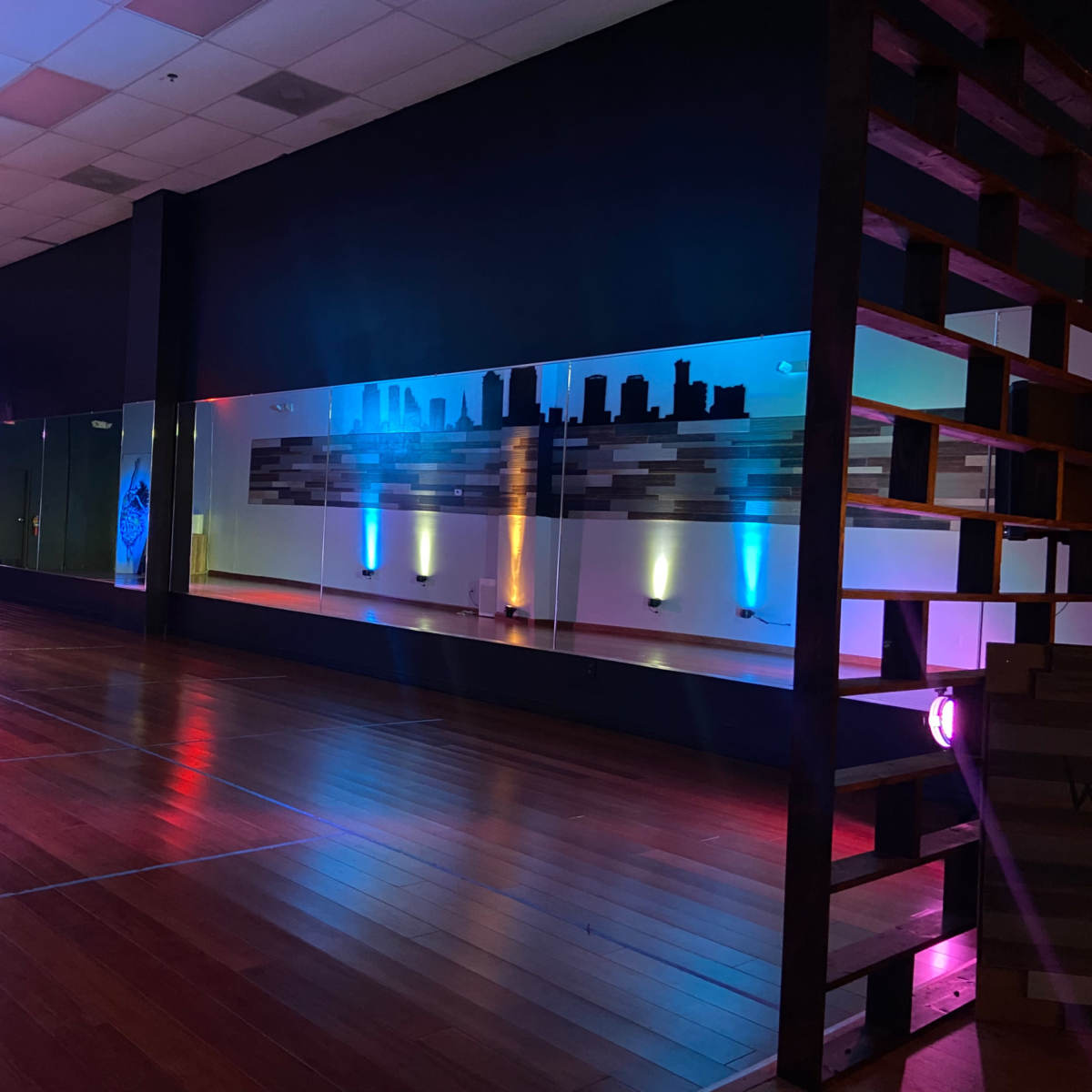 A dance studio with wooden flooring, illuminated by colorful lights and featuring a large mirrored wall that reflects a city skyline design.