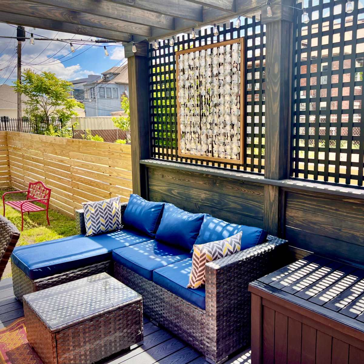 A shaded outdoor patio features a dark wicker seating area with blue cushions, a decorative wall of lattice, and a small coffee table.