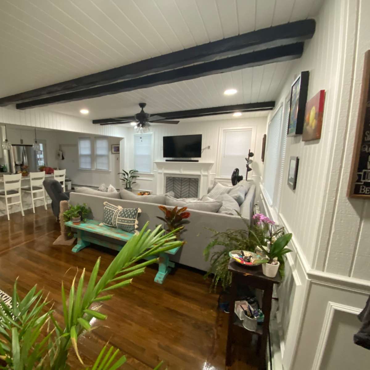 A living room with a grey sofa, wooden beams on the ceiling, a coffee table, and a view of a kitchen area with white furniture in the background.