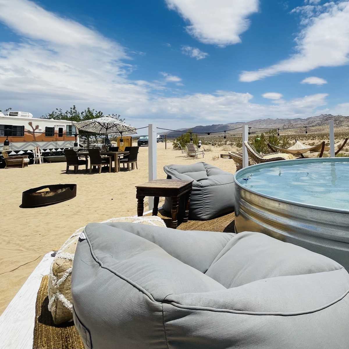 A round metal tub filled with water is set up in a desert landscape, surrounded by large bean bags, a picnic table, and an RV in the background.